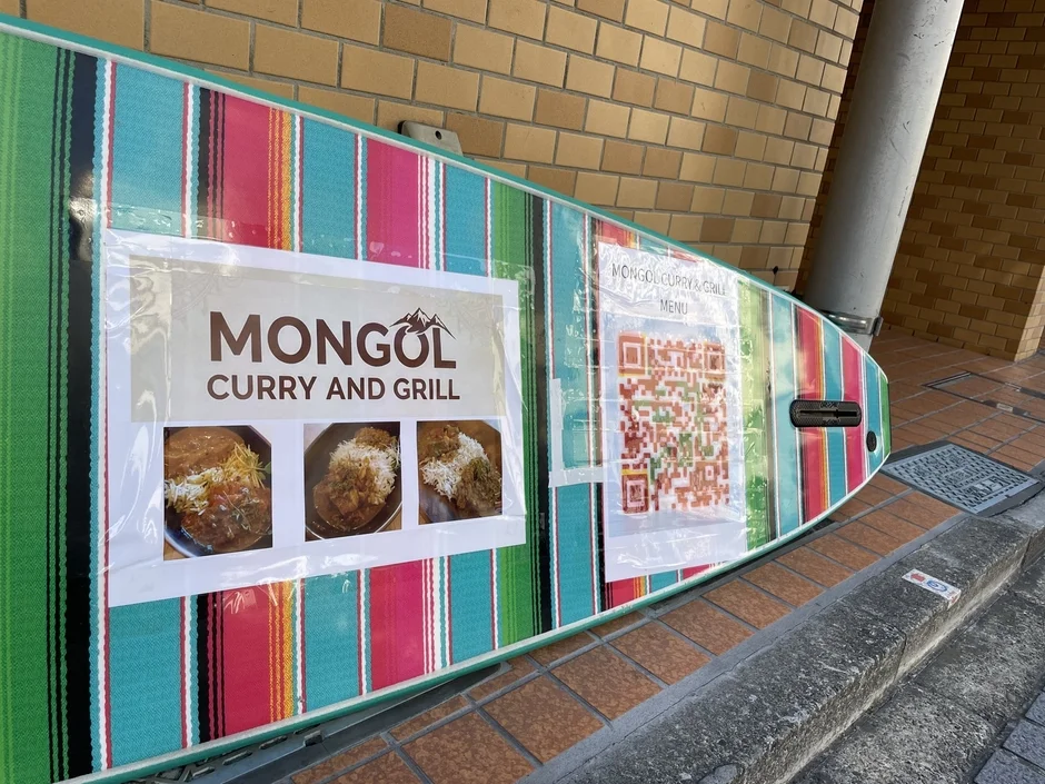 カラフルなストライプ模様のボードに「MONGOL CURRY AND GRILL」の文字と料理の写真、QRコードが貼られた看板。モンゴル料理店のメニューや宣伝として使われているようです。
