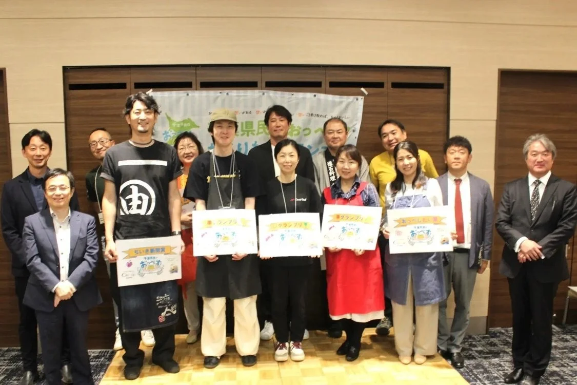 この画像は、多くの人々が集まって、おそらく「県民おやつ」に関連するコンテストかイベントの表彰式で記念撮影をしている様子です。参加者は「グランプリ」や「ちいき新聞賞」と書かれた賞状を手に笑顔を見せています。