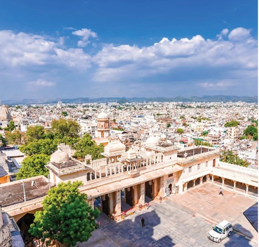 インドの歴史的な都市を上空から捉えたパノラマ写真です。手前には装飾的な宮殿のような建物とその広場があり、遠方には白い建物が密集した市街地が広がっています。青い空には白い雲が浮かび、美しいコントラストを見せています。