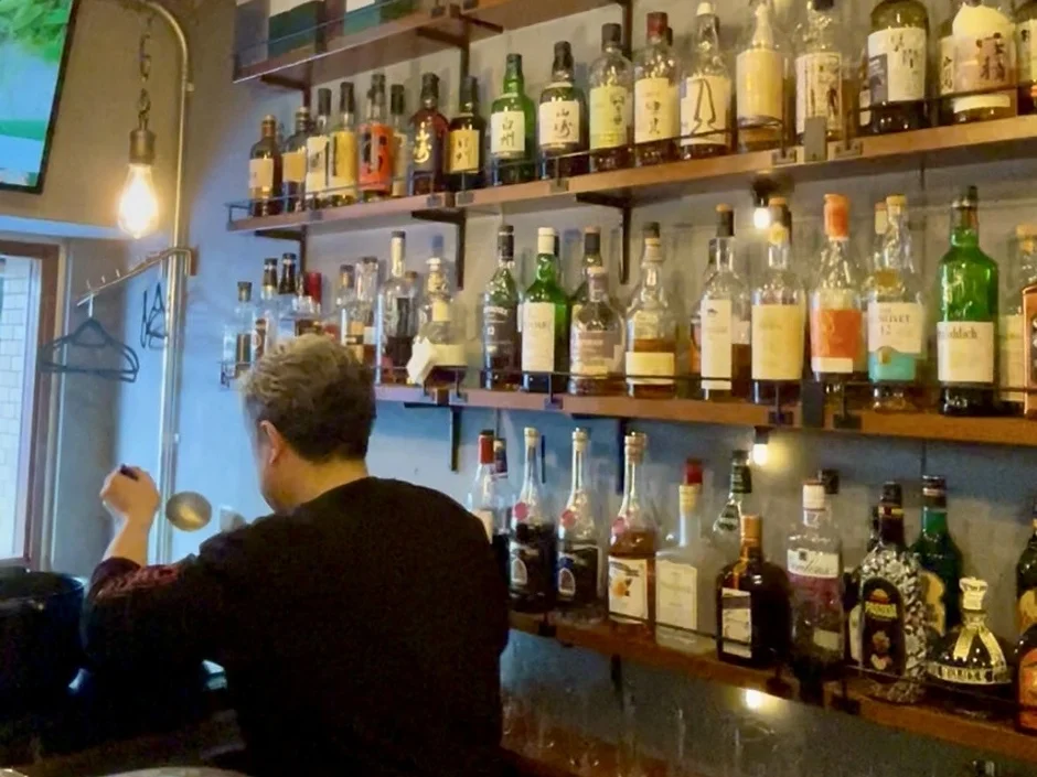 バーカウンターで背中を向けた人物が作業しており、その背後には様々な種類のウイスキーや洋酒のボトルがずらりと並んだ棚が印象的です。暖色系の照明が落ち着いた雰囲気を醸し出しています。