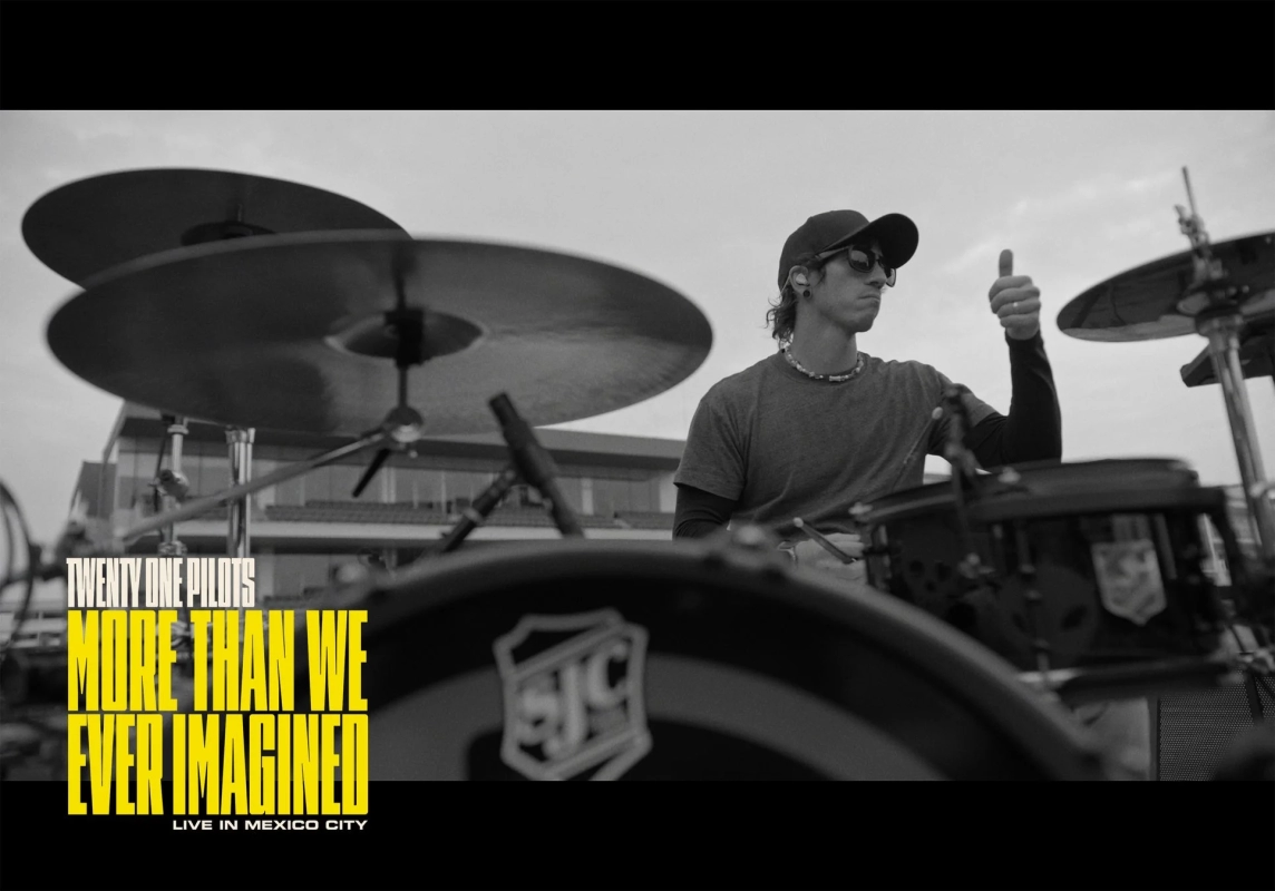 モノクロの画像で、ドラマーが屋外でドラムを演奏し、親指を立ててポーズをとっている