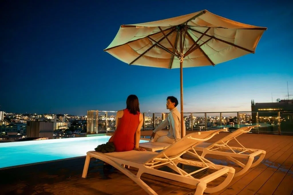夜景が広がる都会のルーフトッププールサイドで、男女がラウンジチェアに座り、リラックスした時間を過ごしています。高級感のある大人の空間です。