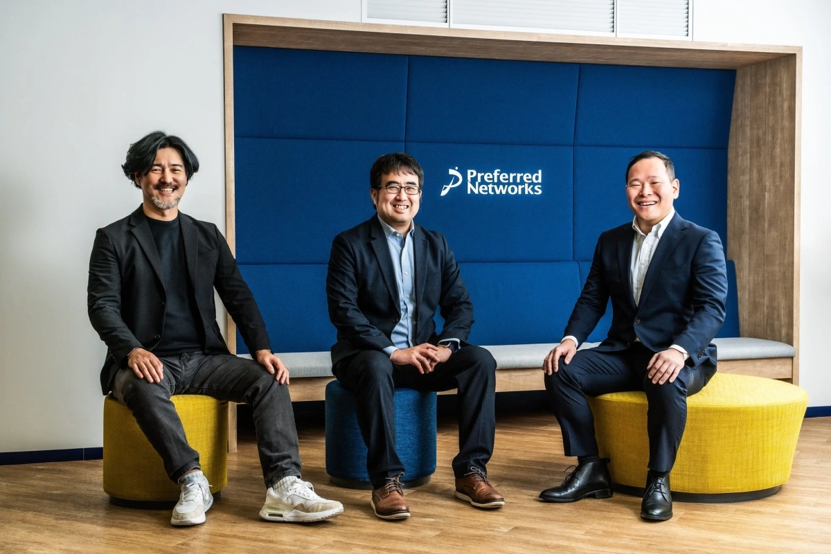 Preferred Networksのロゴがある青い壁を背景に、3人の男性がスツールに座って笑顔でカメラを見ている様子