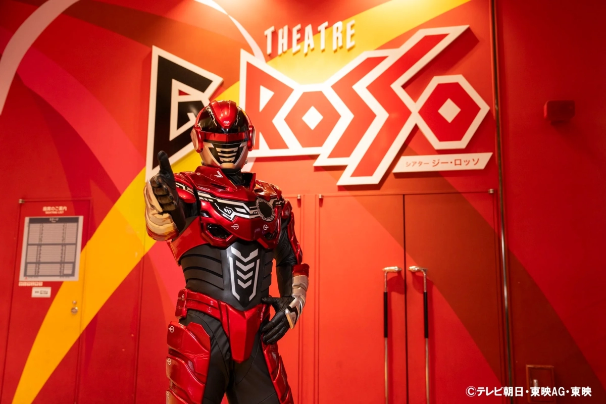 赤いヒーローがGロッソのロゴの前でポーズ