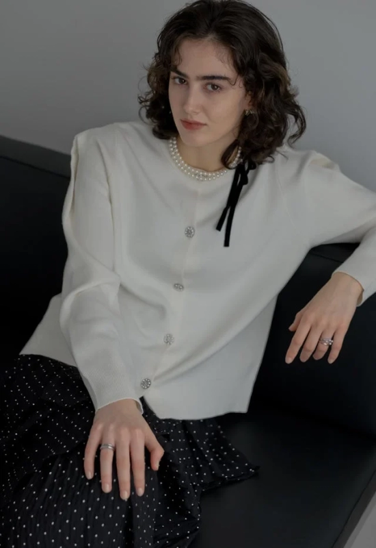 白いカーディガンとドット柄のスカートを着用した女性が、ソファに座ってカメラを見つめている画像です。パールネックレスと黒いリボン、指輪がエレガントな印象を与えています。