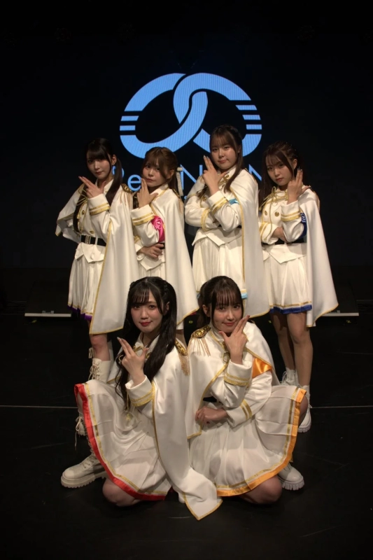 白い衣装を着用した6人の女性アイドルグループが、ステージ上で特徴的なポーズをとって写っている集合写真