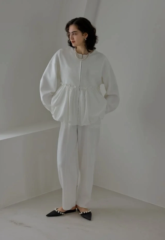 白いセットアップを着た女性が、ミニマルな空間に立っています。ボリュームのあるトップスとワイドパンツに、パールネックレスとパール装飾のフラットシューズを合わせた、清潔感のあるエレガントなコーディネートです。