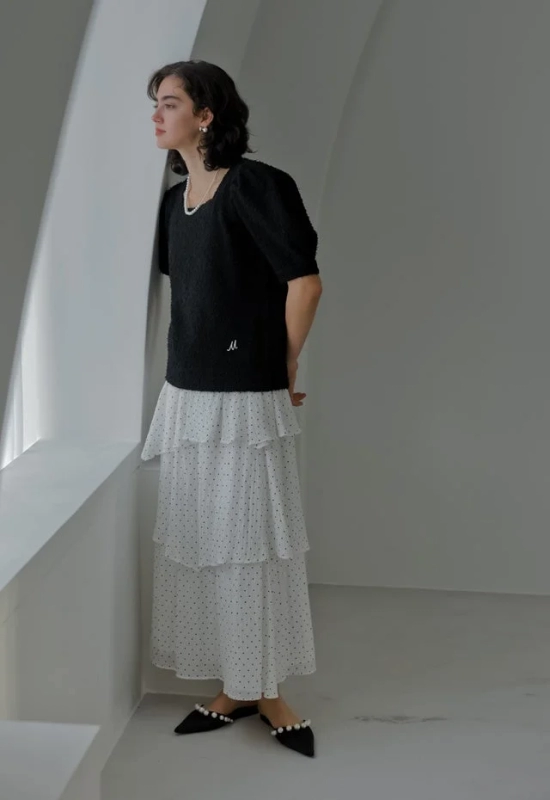 白いティアードのドット柄スカートと黒いパフスリーブのトップスを着用し、パールアクセサリーを身につけた女性が窓辺に佇んでいます。明るい室内で横顔を見せています。