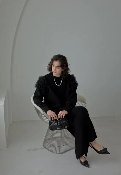 白いミニマルな空間で、黒い服とパールネックレスを身につけた人物がモダンな椅子に座っています。チュールのような袖のデザインが特徴的なトップスと、黒いクラッチバッグ、パンプスを合わせたシックなファッションスタイルです。