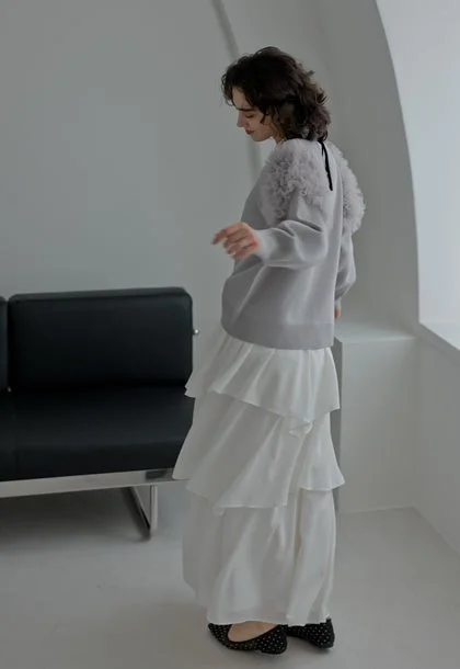 ふわふわの飾りがついたグレーのセーターと白いティアードロングスカートを着用した女性の全身写真。室内でポーズをとっており、フェミニンで洗練されたファッションスタイルが特徴です。