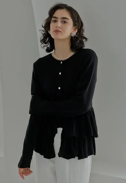 黒いティアードデザインのカーディガンと白いパンツを着用した女性のポートレート。パールネックレスとイヤリングを身につけ、カメラを見つめている。シンプルで洗練された印象のファッション写真。