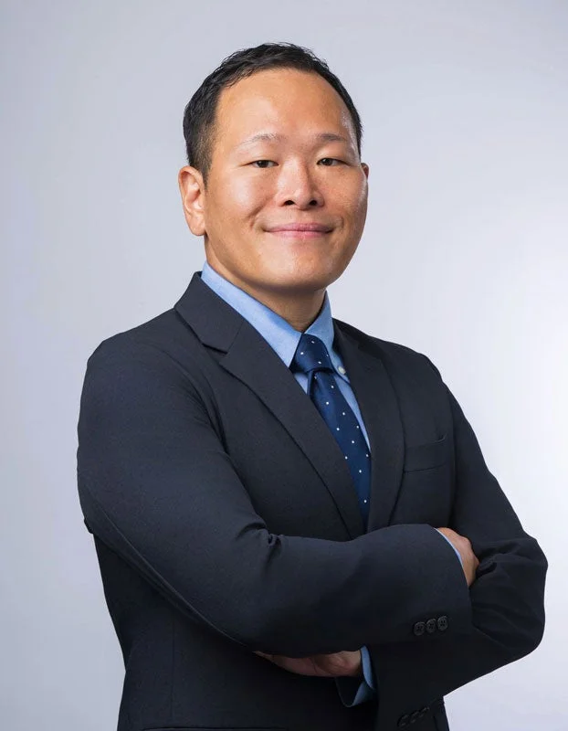 グレーの背景の前で、ダークスーツと水色のシャツ、ネクタイを着用したアジア系の男性が腕を組み、笑顔で立っているポートレート写真