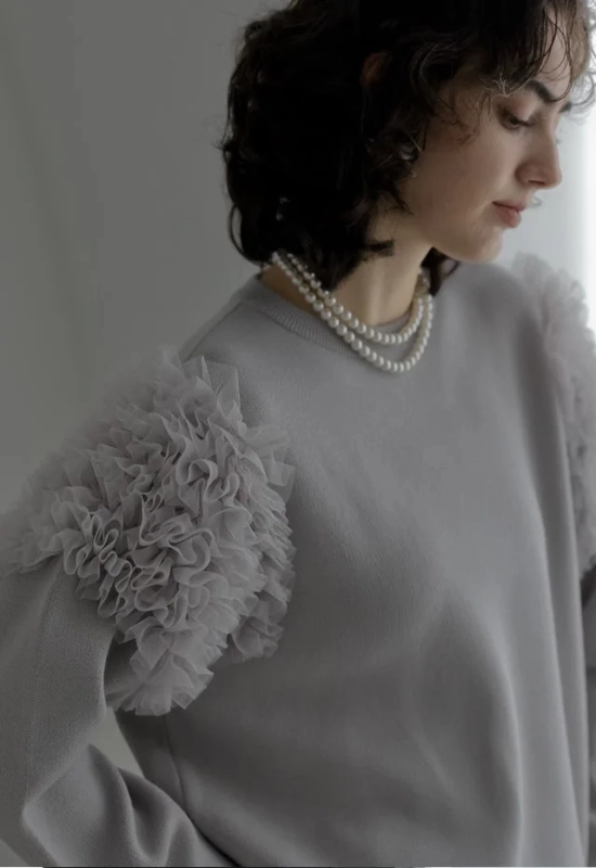 フリル袖が特徴的なライトグレーのセーターを着用し、二連のパールネックレスを合わせた女性のポートレート。短くカールした髪型が上品で洗練された印象を与えています。